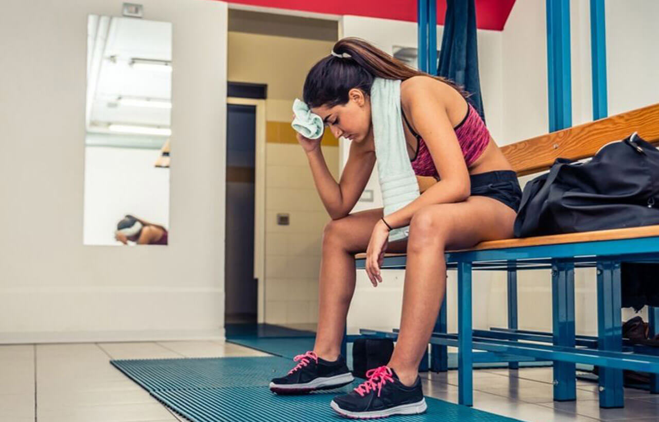 Mulher sentada no vestiário da academia secando o suor do rosto após exercício físico