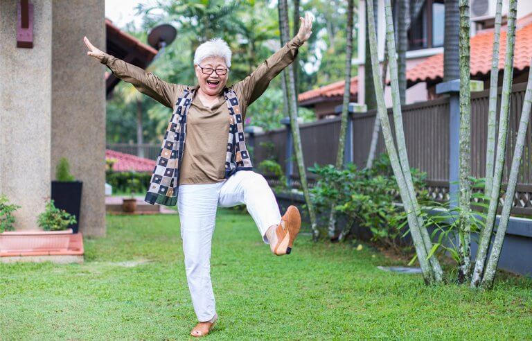 Idosa feliz em um jardim com uma perna para o alto, braços para cima e sorriso no rosto
