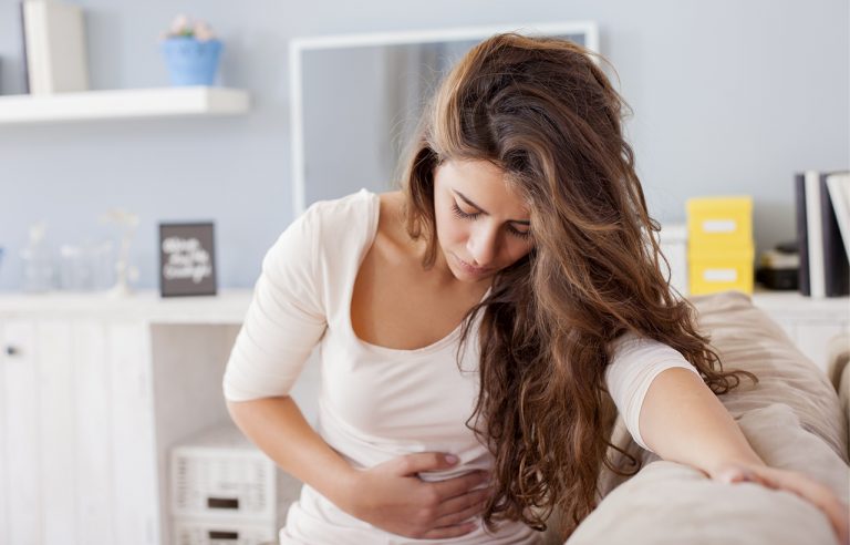 Mulher jovem apoiada em sofá com mãos na barriga e expressando dor ou cólica