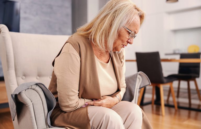 Mulher de cabelo grisalho com mãos na barriga e cabeça baixa por sentir cólica intestinal