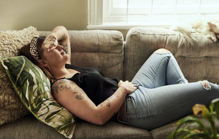 Mulher tatuada deitada no sofá com as mãos na barriga por estar com dor ou cólica