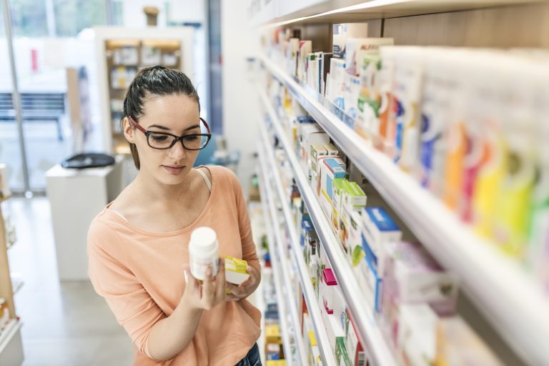 Mulher jovem de óculos comprando remédio na farmácia em frente a prateleira de medicamentos