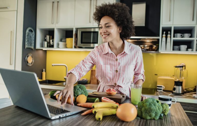 Mulher na cozinha com frutas, legumes, vegetais e com notebook para fazer receita saudável