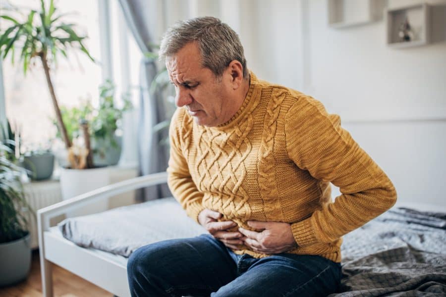 Homem branco sentado na cama utilizando um suéter amarelo com as mãos na barriga e com uma expressão de dor e desconforto
