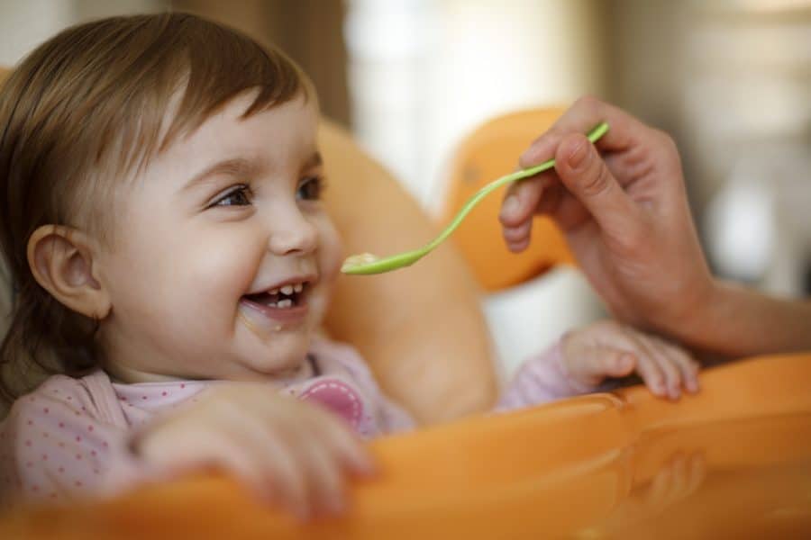 Bebê menina com a pele branca sorrindo enquanto é alimentada pela mãe com uma colher verde